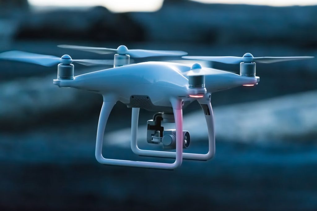 A white quadcopter drone with a mounted camera gimbal hovers in flight against a dim, blurred blue background, its four propellers spinning and red lights glowing near the rotors.