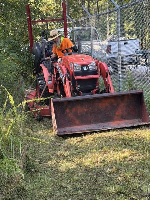 Draughon's Dynamic Enterprise crew member operating a tractor to clear grass and vegetation along a chain-link fence.